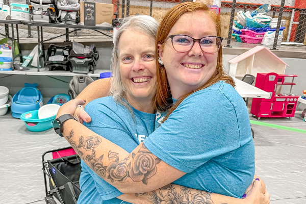 Mom wears baby and holds toddler boy clothes in each hand. Second mom shops behind her from the racks of boys clothes.